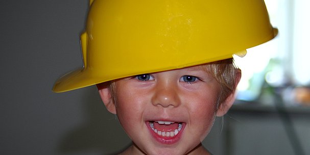 lächelndes Kleinkind, ca. 3 Jahre mit gelben Bauarbeiterhelm auf dem Kopf.